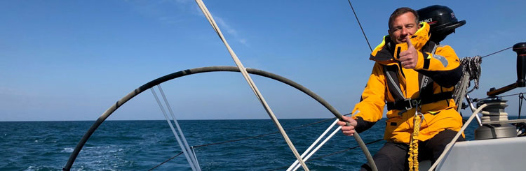 Foto an Bord eines Segelboots von einer Person in orangefarbener Rettungsweste, die das Steuerrad hält und mit erhobenem Daumen in die Kamera schaut
