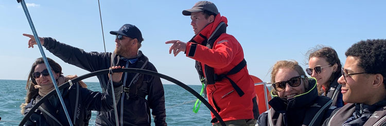 Foto, das in einem Boot von einer Gruppe von Personen aufgenommen wurde, die neben dem Steuerrad sitzen. Zwei stehende Personen zeigen auf den Horizont und eine Person hält das Ruder.